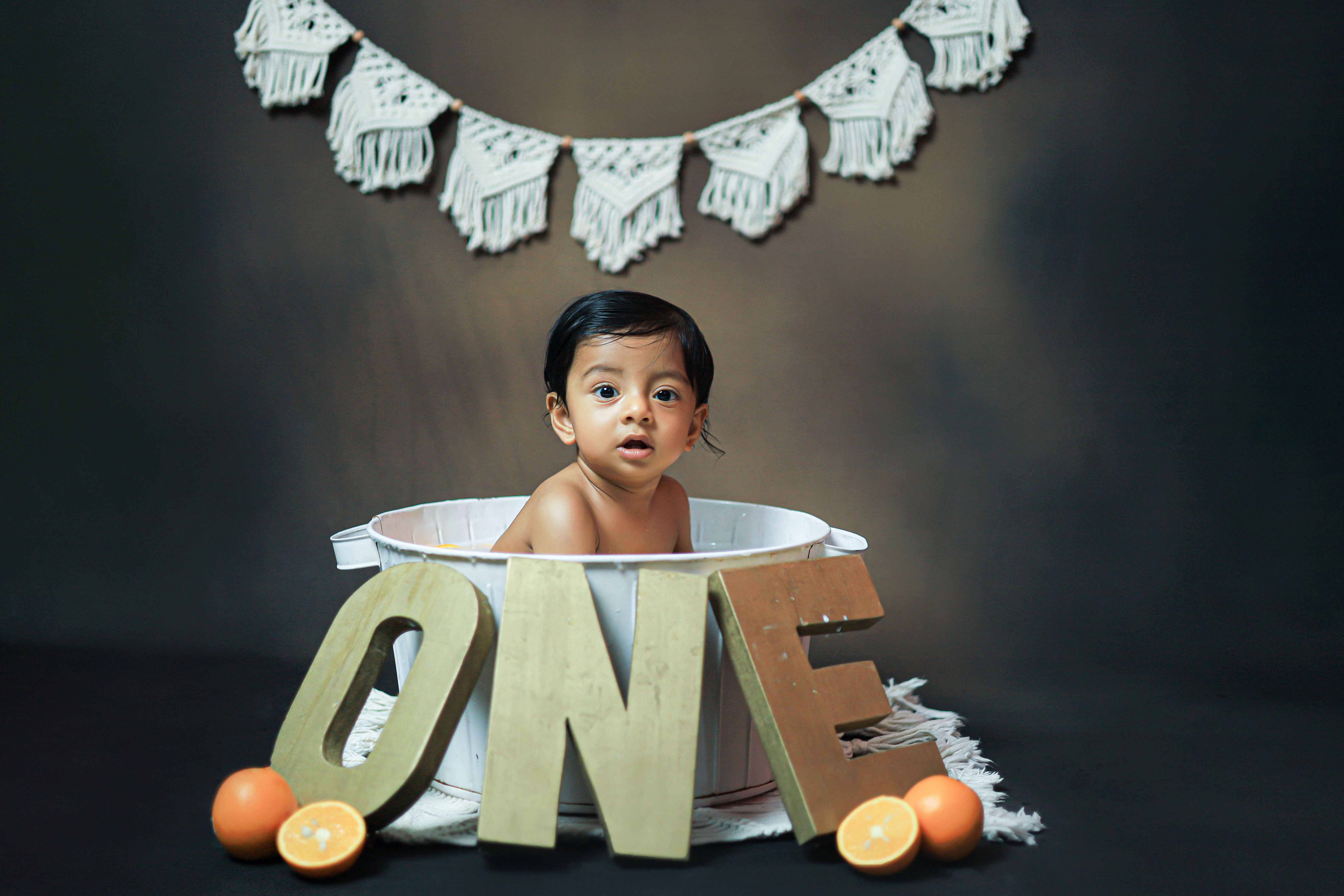 A happy baby in a bath tub with a one-year prop, looking delighted during a beautiful baby photoshoot.
